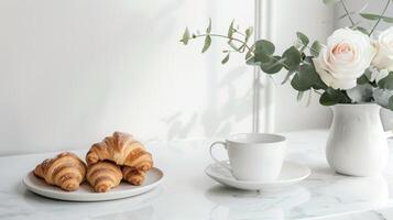 On a white marble table there is a vase with white roses, aromatic coffee in a cup, crispy fresh croissants photo