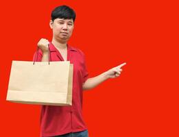 Happy asian man carrying brown shopping bags and point your finger at the empty space. on red background. Chinese New Year theme photo