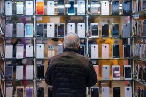 A pensioner stands helplessly in a smartphone store with many new smartphone models. AI generate photo