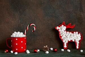 Christmas or New Year greeting card. Red cocoa mug and deer with marshmallows on table wall background, copy space photo