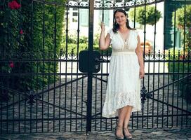 woman in a white lace dress poses in front of a gate photo