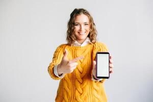 Smiling Woman Pointing at Mobile Phone Screen. Happy woman pointing at mobile phone screen, showcasing a new app. Holding a smartphone with a blank display, isolated on white background. photo