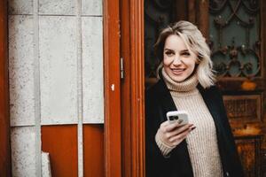 Smiling Woman in Black Coat with Smartphone. Cheerful woman with a black coat and white sweater, holding a smartphone. Ideal for concepts related to digital communication, professional networking photo