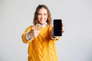 Happy Woman Pointing at Mobile Phone Screen, Smiling young woman pointing at mobile phone screen, presenting a new app. Holding a smartphone with a blank display, isolated on white background. photo