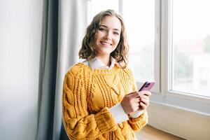 Happy Woman Using Smartphone Indoors. Smiling young woman using her smartphone at home, enjoying online shopping, messaging, and browsing. Capturing the essence of modern lifestyle and technology photo