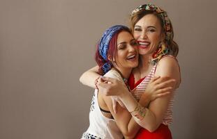 Portrait of two beautiful women in bright clothing and bow in head with bright lips hugging in studio on brown background photo