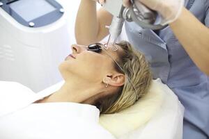 Female cosmetician doing laser resurfacing to client in glasses lying on couch in office photo