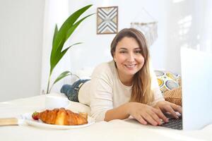Smiling female freelancer working on laptop and having breakfast in bed photo