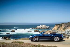 Luxury New convertible parked on scenic coastal road with ocean waves crashing on rocks and clear blue sky as background photo
