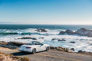 Luxury New convertible parked on scenic coastal road with ocean waves crashing on rocks and clear blue sky as background photo