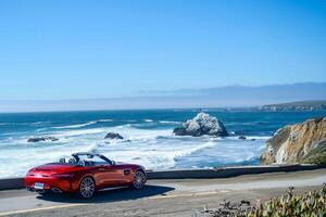 Luxury New convertible parked on scenic coastal road with ocean waves crashing on rocks and clear blue sky as background photo