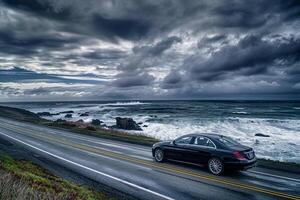 Compact Sedan on Scenic Coastal Highway with Ocean Waves Crashing on Rocks and Dramatic Cloudy Sky Background in Nature photo