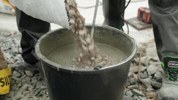 Builder mixing concrete in a black bucket, with the focus on the stirring action. The background consists of gravel, and the person is wearing work pants and sturdy work shoes, indicating a constructi video
