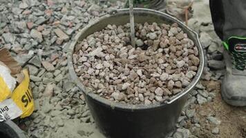Builder mixing concrete in a black bucket, with the focus on the stirring action. The background consists of gravel, and the person is wearing work pants and sturdy work shoes, indicating a constructi video