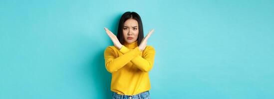 Serious and assertive korean woman showing cross stop gesture, frowning and telling no, prohibit action, disapprove something bad, standing over blue background photo