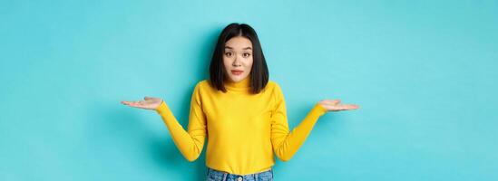 Image of indecisive asian woman shrugging shoulders, spread hands sideways and looking clueless at camera, standing indecisive against blue background photo