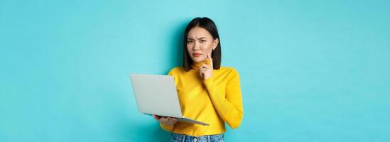Serious looking asian woman working on laptop and thinking, frowning at camera, solving problem at work, standing over blue background photo