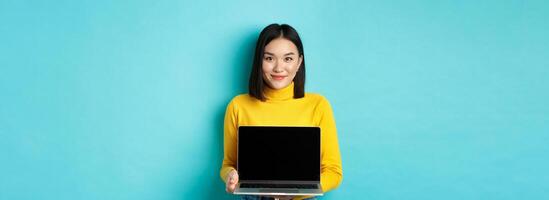 Beautiful and stylish asian woman demonstrate product on screen, showing empty laptop display and smiling, standing over blue background photo