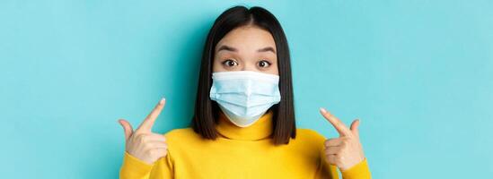 Covid-19, social distancing and pandemic concept. Close up of surprised asian girl pointing at her medical mask and looking amazed at camera, blue background photo