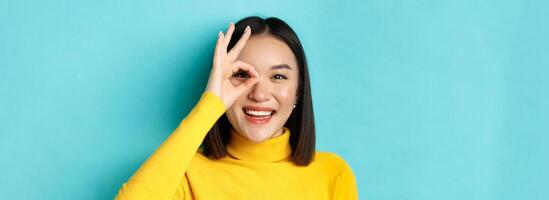 Beauty and makeup concept. Close up of carefree asian girl showing OK sign on eye and smiling, looking happy at camera, standing over blue background photo