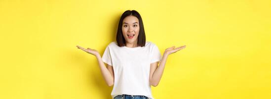 Beauty and fashion concept. Surprised asian girl spread hands sideways, holding on palms, pointing at two products, standing over yellow background photo