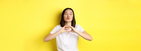Beautiful asian woman showing I love you heart gesture, smiling at camera, standing against yellow background. Concept of valentines day and romance photo