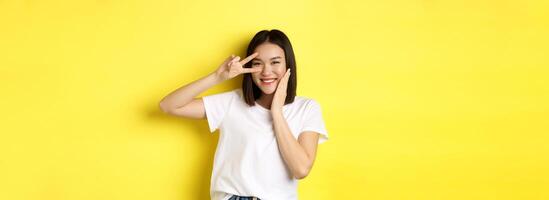 Lovely asian girl in white t-shirt posing with hand on cheek, showing peace sign on eye, standing over yellow background photo