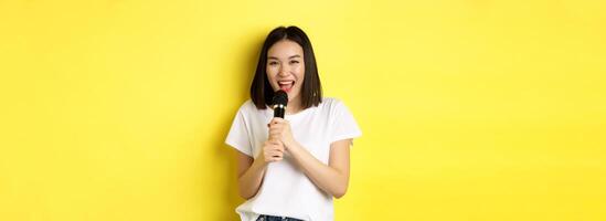 Beautiful asian woman singing karaoke, perform with microphone, smiling happy and looking at camera, standing over yellow background photo