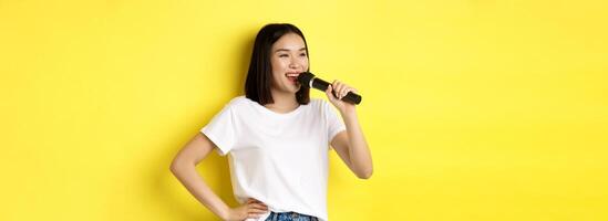 Happy asian woman singing song in karaoke, holding microphone and looking aside with cheerful smile, standing over yellow background photo