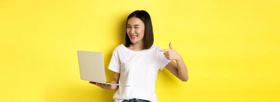 E-commerce and shopping concept. Good-looking asian girl winking at camera, smiling and pointing finger a laptop, showing something, standing over yellow background photo