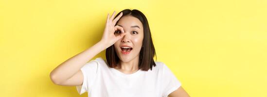 Close up of cute asian girl feeling happy, showing OK sign on eye and smiling, standing over yellow background photo