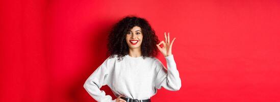 Confident smiling woman in casual clothes, showing OK sign to approve or like product, guarantee good quality, making a compliment, red background photo