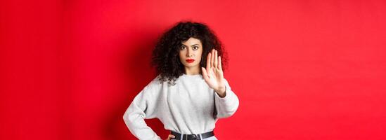 Confident tensed woman extend hand to say stop, disapprove action and prohibit it, make no gesture, standing on red background and forbid something photo