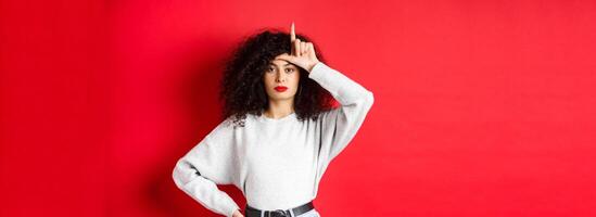Confident european woman with curly hair, mocking lost team, showing loser gesture and staring unimpressed at camera, standing in casual clothes on red background photo