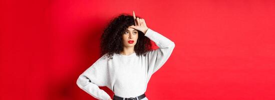 Arrogant woman with curly hair, showing loser sign on forehead and looking aside, mocking someone, standing on red background photo