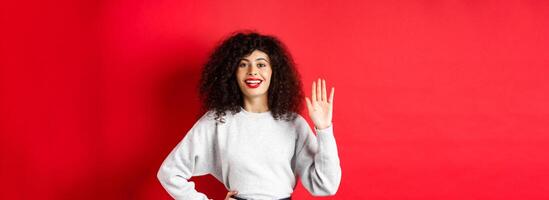 Friendly woman with curly hair greet you, waiving hand and saying hello, introduce herself, standing on red background photo