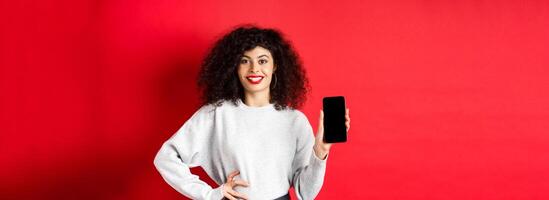 Attractive smiling woman showing empty smartphone screen and looking happy, advertising online store or application, standing against red background photo