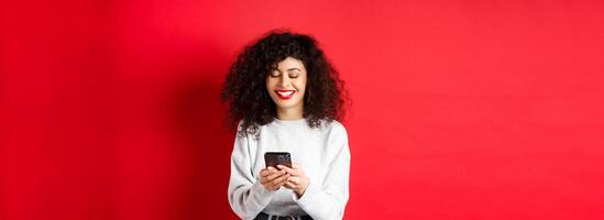 Happy positive girl chatting on phone, reading message and smiling, using social media app, standing on red background photo