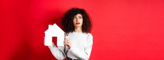 Real estate. Sad woman thinking of buying own house, showing paper home cutout and looking up with uspet face, standing on red background photo