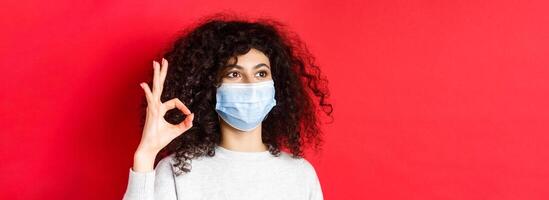 Covid and health concept. Image of young woman in medical mask approve promo, showing okay gesture and looking at empty space, standing on red background photo