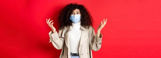 Covid-19, pandemic and quarantine concept. Shocked lady with curly hair and medical mask, raising hands up confused, standing on red background photo