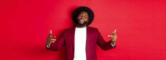Christmas shopping and people concept. Confident and sassy man showing bring it on gesture, dancing hip hop and looking self-assured at camera, red background photo