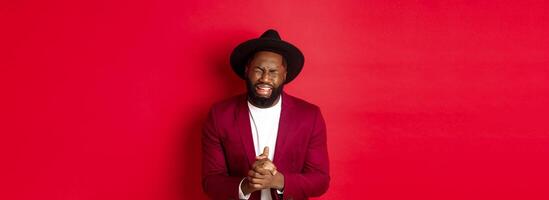 Miserable and heartbroken Black man crying, begging for help, holding hands in pray, standing against red background photo
