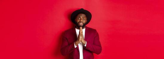 Miserable Black man asking for help, begging for favour and looking sad, standing against red background photo