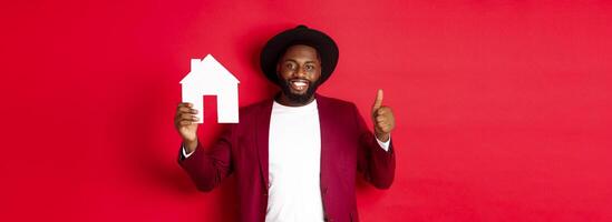 Real estate. Stylish african american bearded man showing home maket, thumb up, recommending agency or apartment complex, standing over red background photo