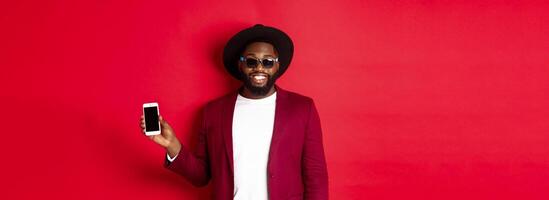 Handsome and stylish Black man showing phone screen at camera, recommending online store or application, standing over red background photo