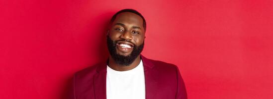 Close-up of handsome bearded Black man celebrating New Year, wearing party outfit and smiling happy, standing over red background photo