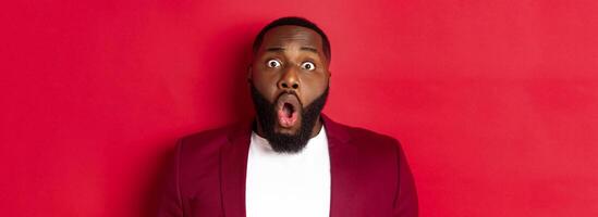 Close-up of shocked Black man gasping and dropping jaw impressed, staring at camera, standing over red background photo
