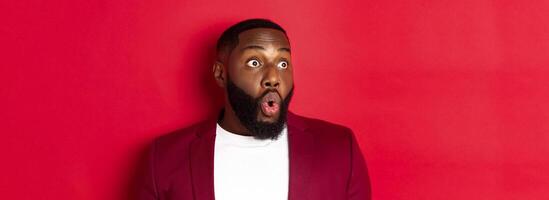 Close-up of impressed Black man looking left at copy space, saying wow and staring with amazement, standing over red background photo