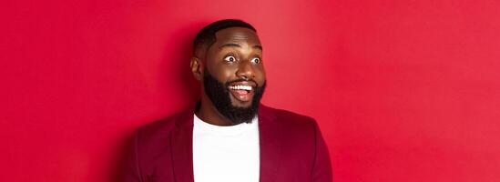 Close-up of excited african american guy looking left, smiling with amazement and joy, standing in blazer against red background photo